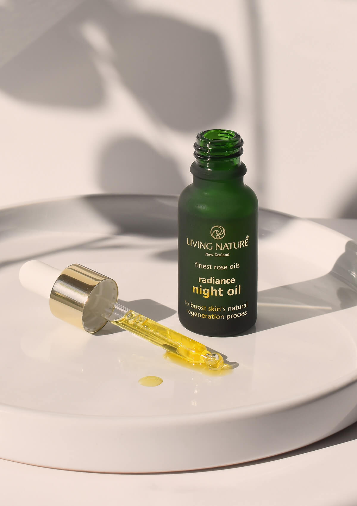 auf einem weißen runden Tablett steht eine offene grüne Glasflasche des Antifaltenrosenöls. Davor liegt eine befüllte Pipette mit dem goldenem Öl