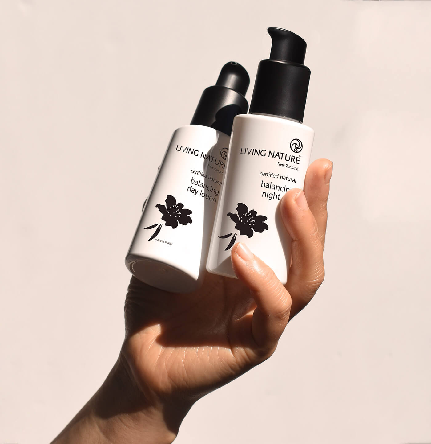 Frauenhand hält zwei weiße Flaschen, Living Nature Balancing Day Lotion und Balancing Night Lotion mit schwarzen Pumpspenderkopf in der Hand und hält sie hoch.