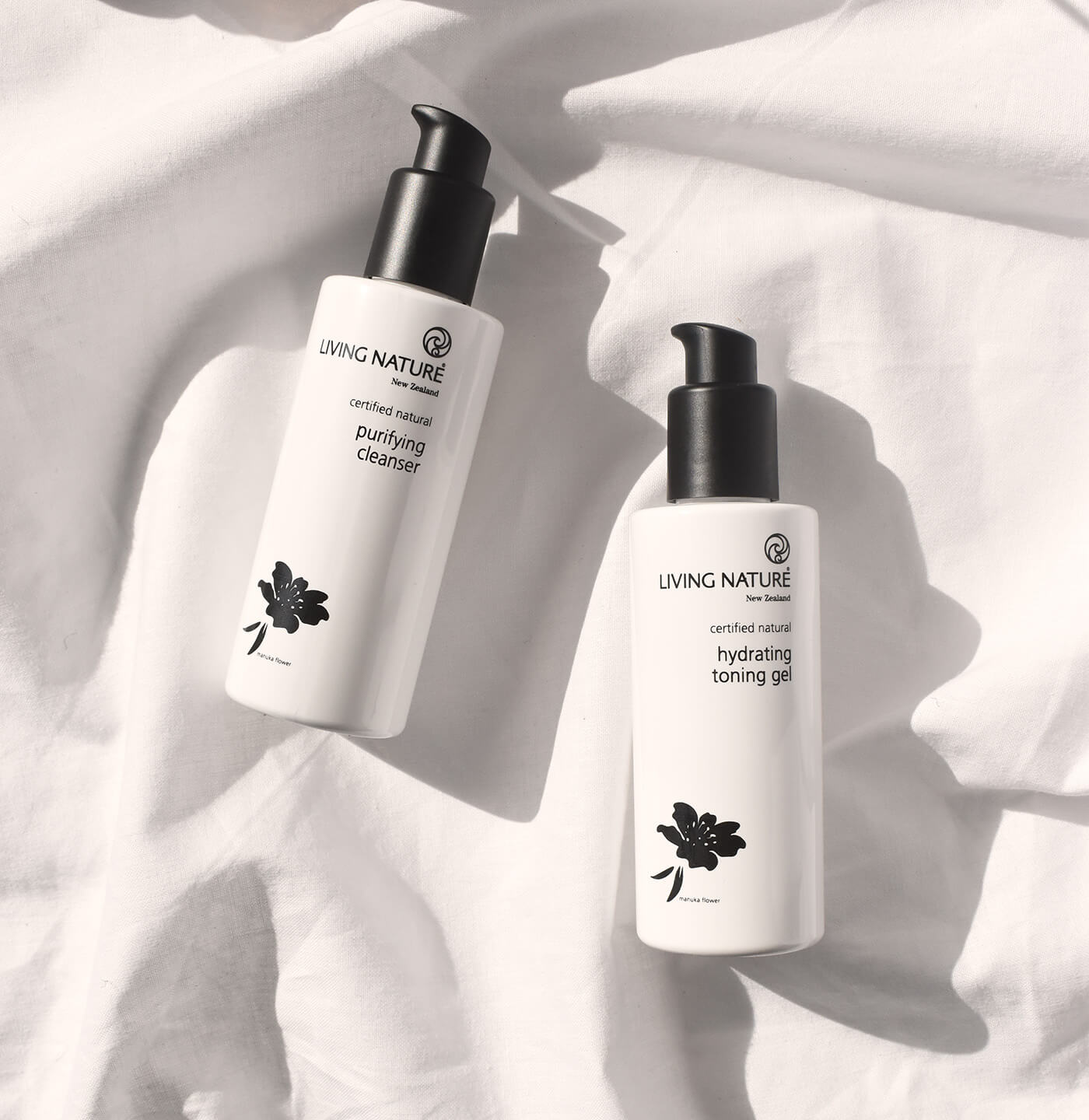 Zwei weiße Flaschen mit schwarzem Pumpspenderkopf vom Living Nature Purifying Cleanser und Hydrating Toning Gel liegen auf einem weißen Leinentuch. Helle, strahlende Anmutung durch Sonnenlicht.