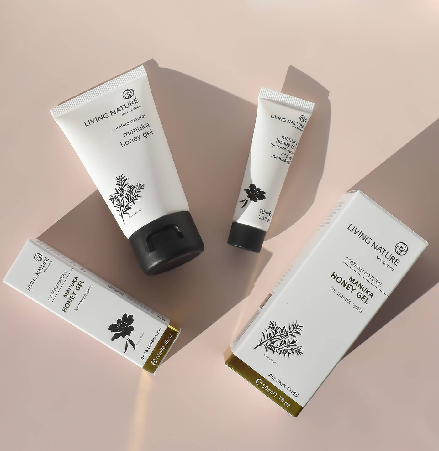 Eine große und eine kleine weiße Tube vom Living Nature Manuka Honey Gel mit schwarzem Deckel liegen auf einem hellrosa Hintergrund mit langen Schatten vom Sonnenlicht. Daneben liegt jeweil die passende Verpackung, weiße Schachtel mit schwarzer Blume als Grafik und goldener Rand.